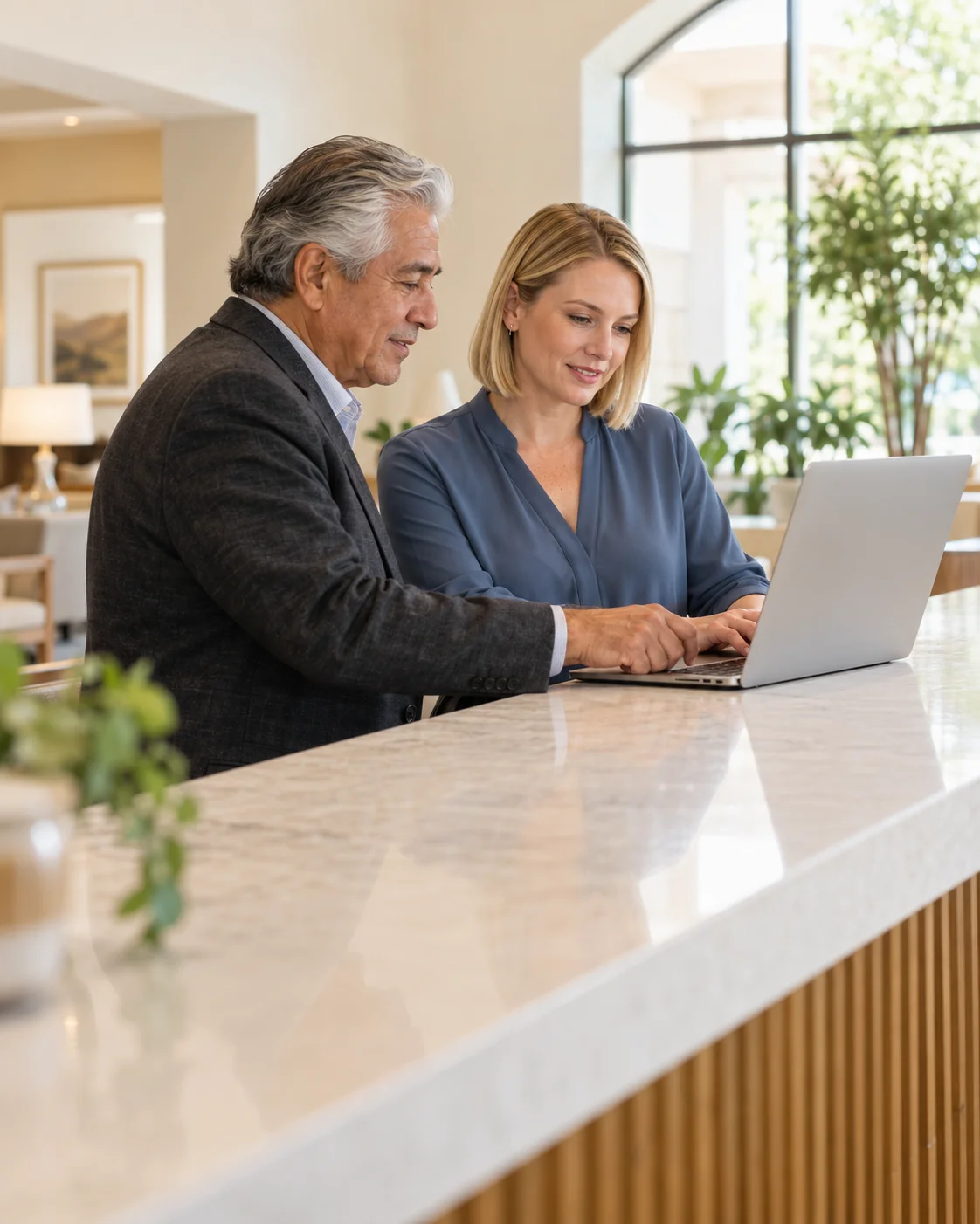 A community director and a sales lead review a laptop together at a long sunlit reception counter, side view with the screen turned away