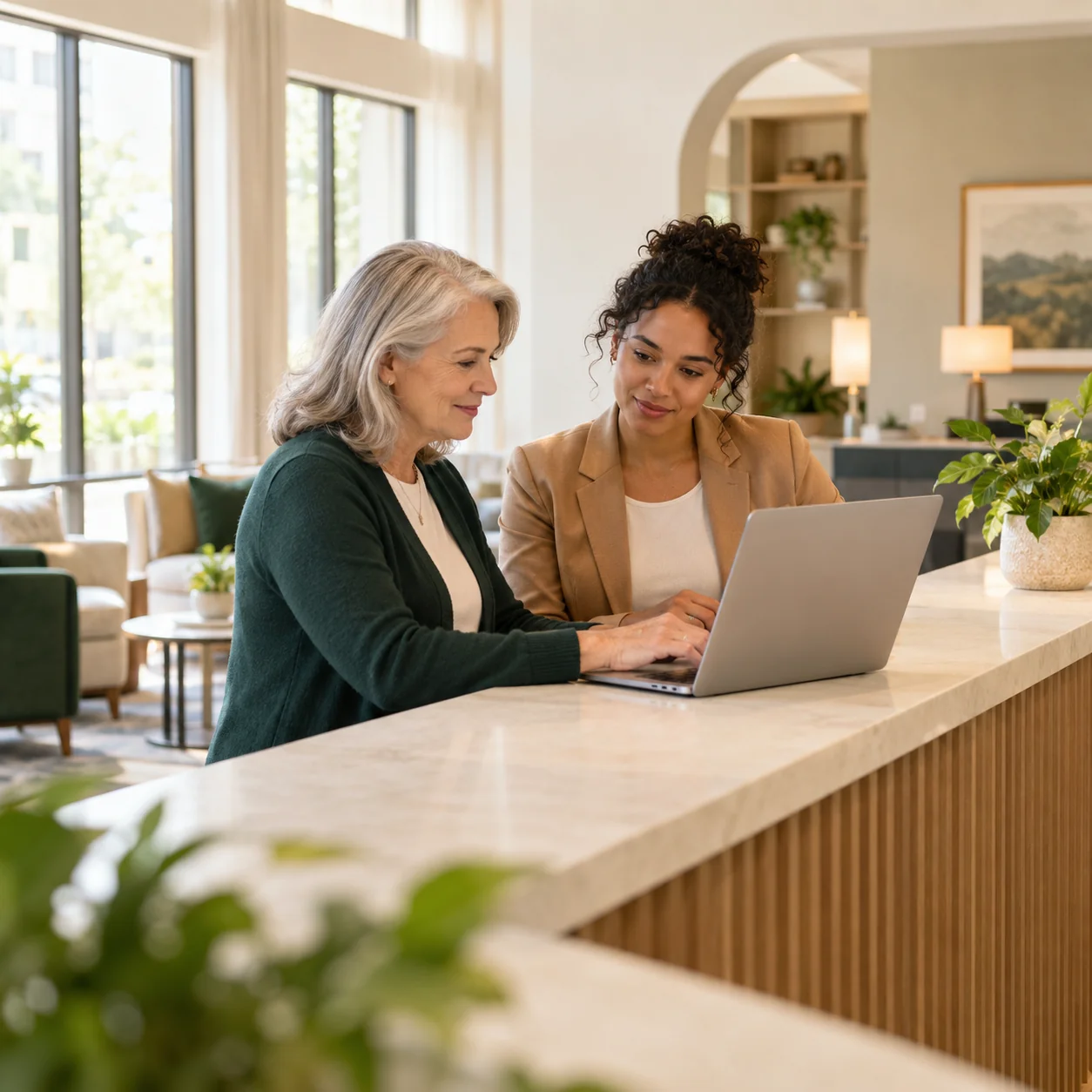 A community director and a sales lead review a laptop together at a long sunlit reception counter, side view with the screen turned away