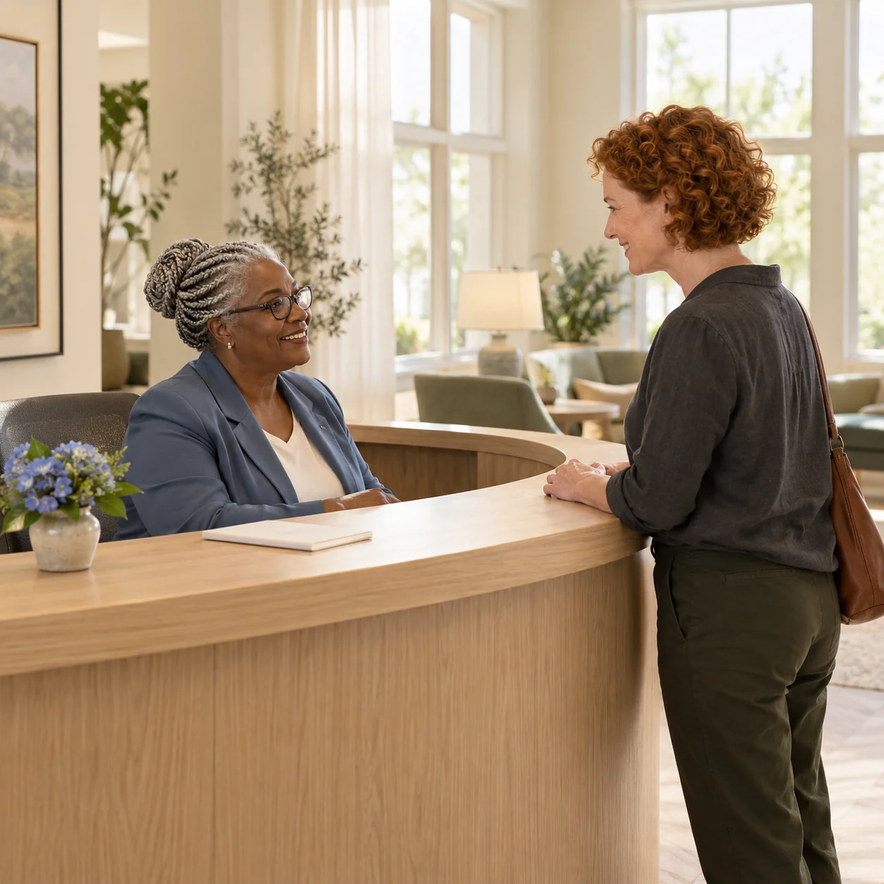 A receptionist welcomes an adult daughter at a curved oak front desk, a plain notepad and a small vase nearby