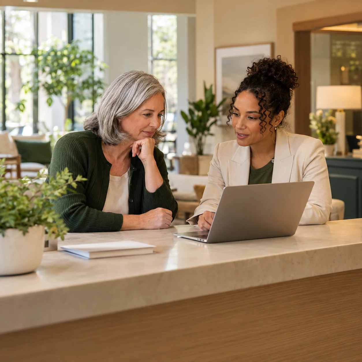 A community director and a sales lead review a laptop together at a long sunlit reception counter, side view with the screen turned away