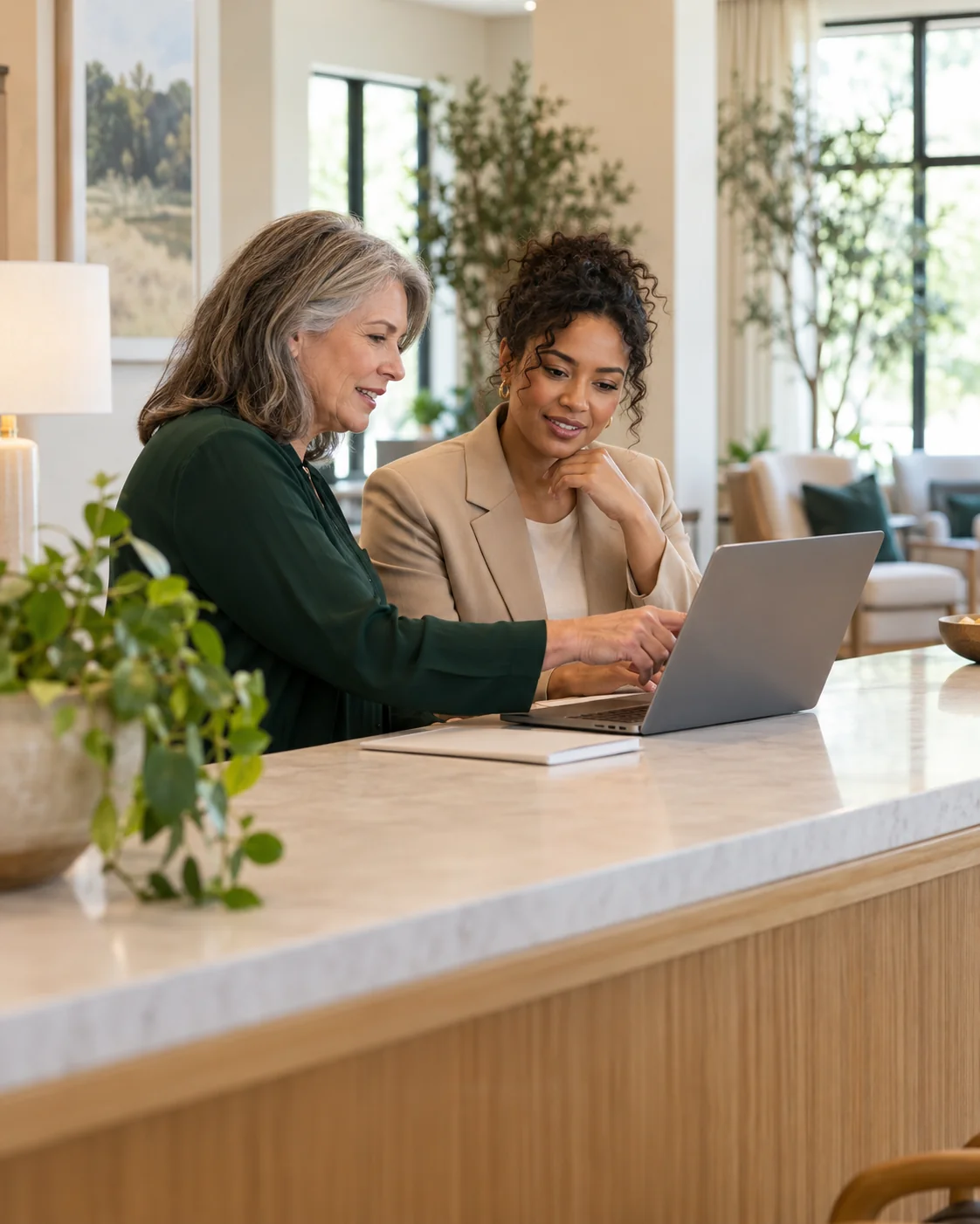 A community director and a sales lead review a laptop together at a long sunlit reception counter, side view with the screen turned away